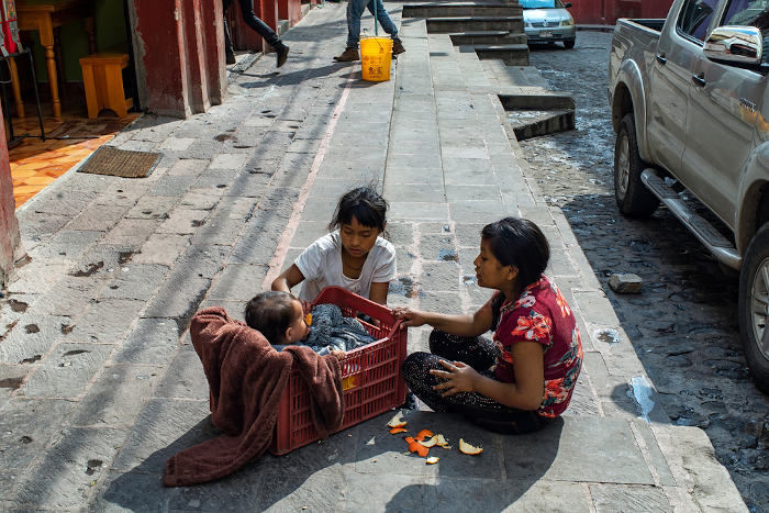 Duas crianças, sentadas na rua, alimentando um bebê, que está dentro de uma caixa
