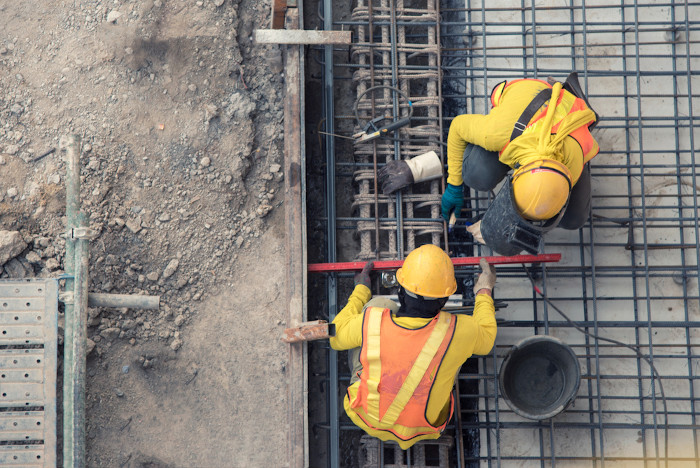 Vista aérea de dois trabalhadores da construção civil, com equipamentos chamativos de proteção, no canteiro de obras