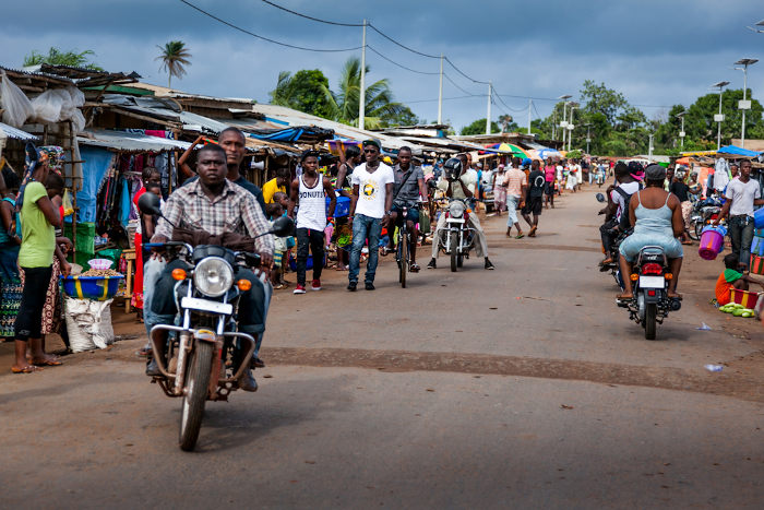 Rua com barracas de feira em suas margens e sendo perpassada por transeuntes, em Serra Leoa 