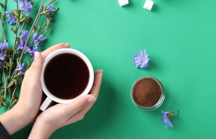 Mãos segurando xícara com chá de almeirão, recipiente com café, flores lilás e cubos de açúcar sobre fundo verde-água.