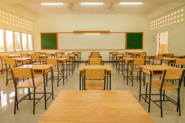Vista do fundo de uma sala de aula, com carteiras em primeiro plano e quadro-negro em segundo plano