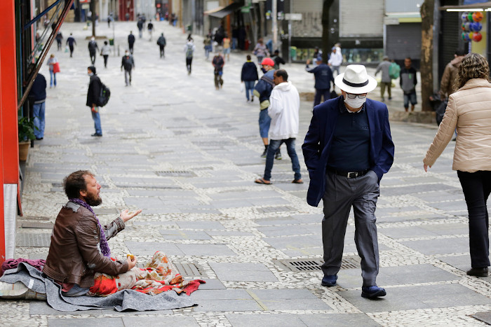  Homem sentado pedindo esmola na rua enquanto pessoas passam por ele.