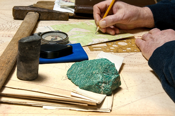 Vista aproximada de um geólogo trabalhando em uma mesa com mapas, martelo, bússola, lupa e uma amostra de rocha.