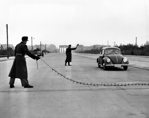 Polícia alfandegária de Berlim Oriental, no contexto da Guerra Fria, parando um carro com uma pesada corrente farpada destinada a furar pneus.