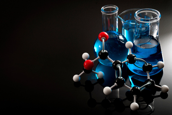 Vista aproximada de frascos de ensaio com líquido azul próximo a uma molécula de benzoato sobre uma mesa.