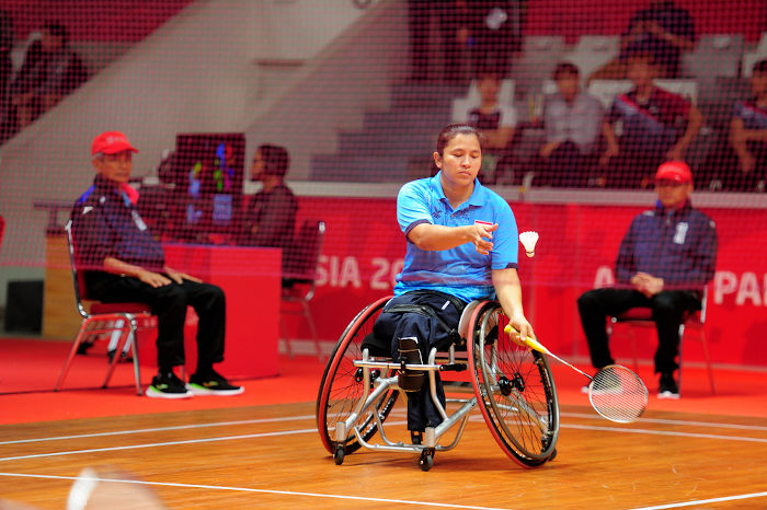 Atleta em cadeira de rodas com camiseta azul prepara raquete para realizar serviço, golpe na peteca (saque). [3]