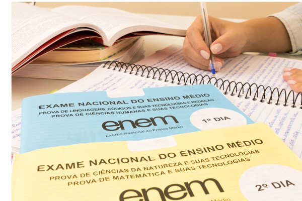 Cadernos de prova do Enem próximos a uma pessoa escrevendo uma redação, avaliada no exame com base em cinco competências.