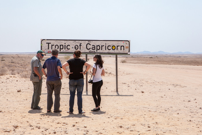 Turistas na Namíbia olhando uma placa indicando que o Trópico de Capricórnio corta a região.