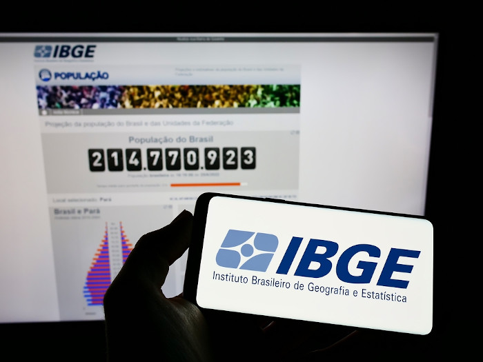 Celular com logo do IBGE em frente a um computador aberto no site do IBGE, representando os dados do censo demográfico.