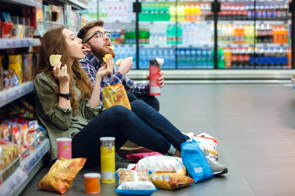 Homem e mulher sentados no chão de um supermercado comendo batatas industrializadas, um tipo de alimento ultraprocessado.