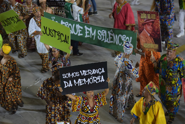 Integrantes de uma escola de samba vestidos com fantasias de Carnaval e segurando cartazes durante desfile.
