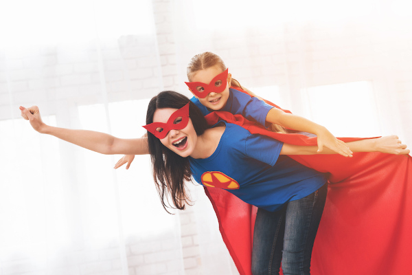 Crianças nas costas de uma mulher e ambas estão vestidas de super-heroínas.