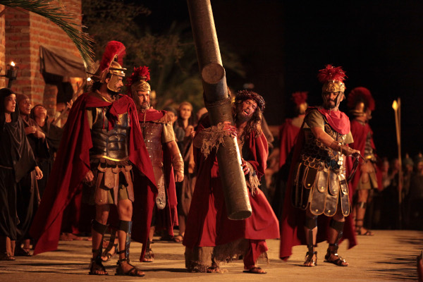 Homem segurando uma grande cruz de madeira, cercado por dois soldados romanos em peça teatral comum na Sexta-Feira Santa.