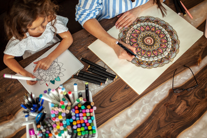 Mulher e criança colorindo mandalas sobre uma mesa de madeira como representação do uso da mandala na educação.
