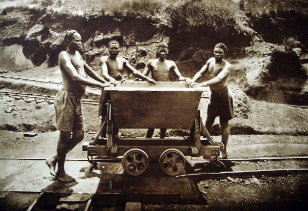 Quatro homens congoleses segurando um carrinho nos trilhos de uma mina.