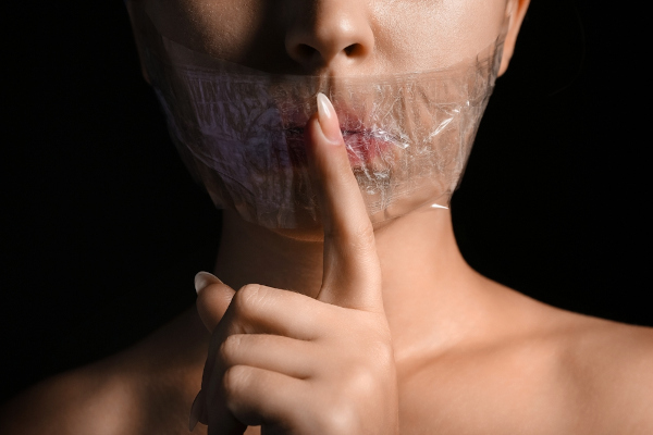 Mulher com mordaça sobre a boca e fazendo gesto de silêncio em referência ao conceito de censura.