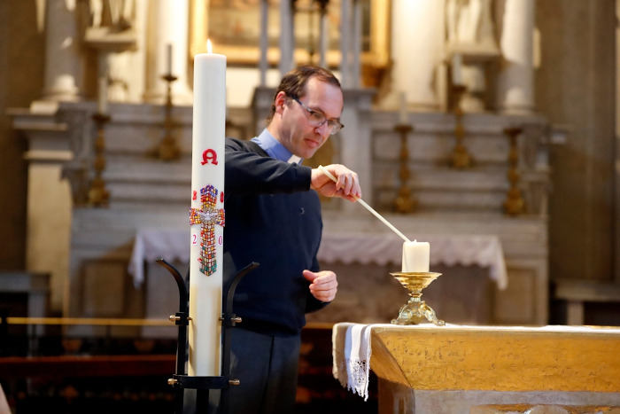 Fotografia de padre acendendo uma vela, em segundo plano; e no primeiro plano, um Círio Pascal, um dos símbolos da Páscoa.