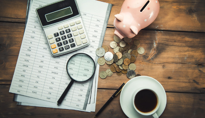 Calculadora, planilhas, lupa, cofre e moedas próximos à xícara de café em alusão ao Imposto de Renda Pessoa Física (IRPF).