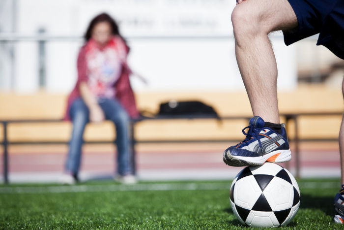 Mulher sentada avistando o pé de um homem apoiado sobre bola de futebol, em alusão ao sexismo.