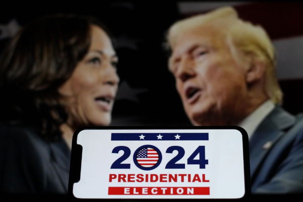 Rostos de Kamala Harris e Donald Trump desfocados. Na frente, um smartphone com o texto: “2024 Presidential election”.