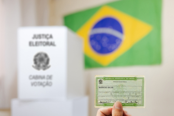 Mão segura título de eleitor; ao fundo, urna eleitoral e bandeira do Brasil.