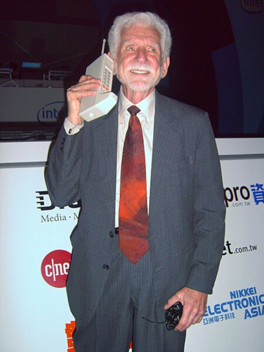 Martin Cooper segurando o protótipo de um celular em texto sobre história do telefone. 