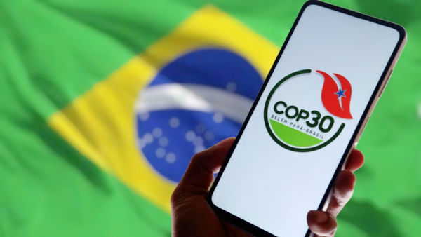 Mão segurando celular com a logo da COP 30 na tela; ao fundo, uma bandeira do Brasil. Mão segurando celular com a logo da COP 30 na tela; ao fundo, uma bandeira do Brasil.