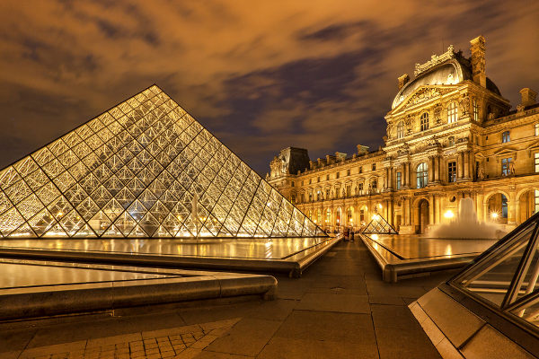 Museu do Louvre à noite. A estrutura em pirâmide de vidros e o prédio ao lado estão iluminados.