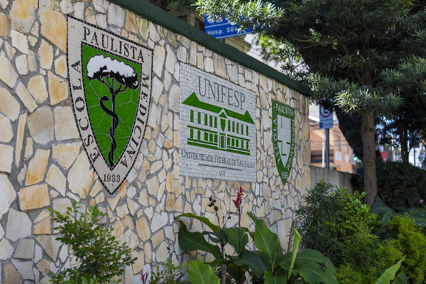 Muro com brasão e logomarca da Unifesp, à frente plantas na cor verde.