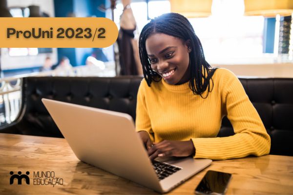 Estudante negra com blusa amarela sorrindo em frente ao computador Texto ProUni 2023/2